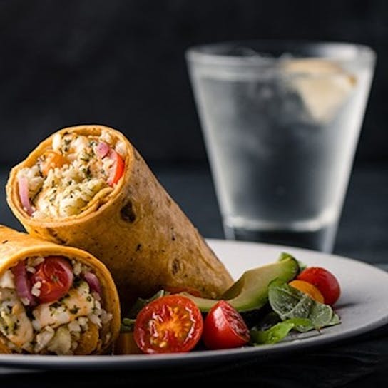 Two stuffed wraps filled with vegetables and herbs are served on a white plate with sliced cherry tomatoes and greens, with a glass of water and ice in the background.