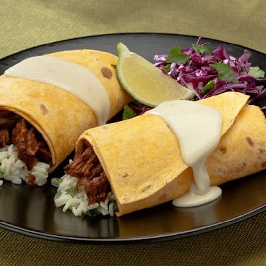 Two burritos filled with shredded meat and rice, topped with white sauce, are on a black plate. The plate also has a wedge of lime, chopped cilantro, and shredded purple cabbage on the side.