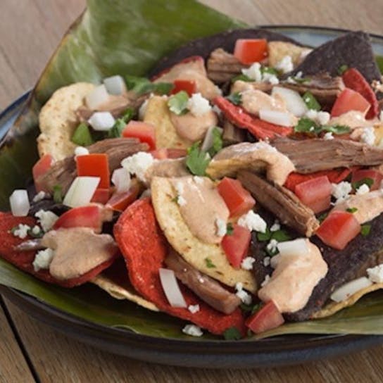 A plate of colorful tortilla chips topped with shredded meat, diced tomatoes, onions, green herbs, cheese, and dollops of sauce, served on a large green leaf on a wooden table.