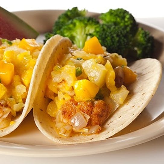 Two soft tacos filled with fish and mango salsa on a plate, served with broccoli florets and two slices of watermelon.