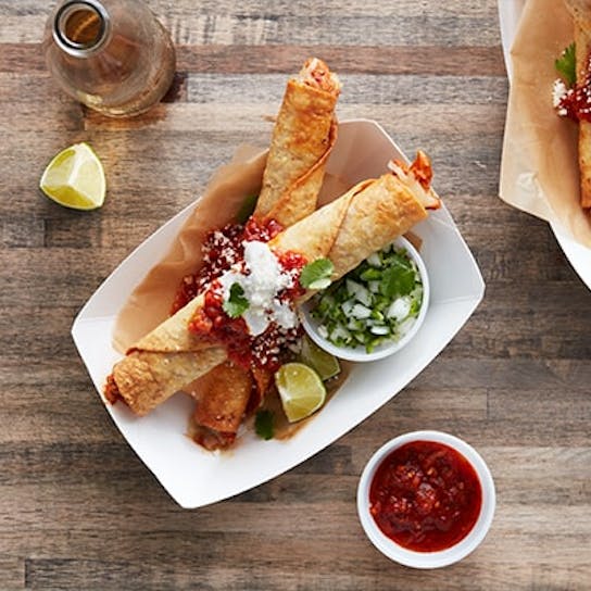Two trays of crispy rolled taquitos topped with salsa and cheese, served with lime wedges, chopped onions, cilantro, and a small bowl of salsa on a wooden table. A drink bottle is partly visible in the corner.
