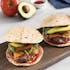 Three burgers with tortilla buns, cheddar cheese, lettuce, tomato, and avocado slices are arranged on a wooden board. An avocado, a tomato, and a small bowl of sauce are in the background.